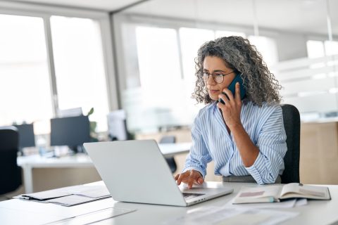 Woman with gray hair talking on a phone while working on a laptop